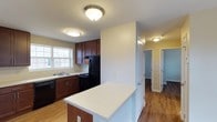 This single-floor space is designed with a functional and cohesive layout, highlighted by warm hardwood-style flooring that flows throughout. The kitchen serves as a central feature, showcasing rich dark wood cabinetry with brushed metal hardware, offering ample upper and lower storage. Black appliances, including a refrigerator, range, and dishwasher, complement the cabinetry, while a double-basin sink is positioned beneath a window that allows natural light to brighten the space. A peninsula with a light-toned countertop extends into the room, providing additional workspace and a natural division between the kitchen and the rest of the living area. Flush-mount ceiling light fixtures illuminate the space with a clean, understated look. The walls are painted in neutral tones, contributing to an open and airy atmosphere. A hallway visible from the kitchen leads to additional rooms, hinting at a practical and well-organized floor plan. The overall design balances utility and comfort, making it a welcoming and move-in-ready space suited for everyday living.