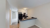 This single-floor residence offers a practical and well-organized layout designed for comfortable everyday living. The kitchen serves as a central feature, showcasing crisp white upper cabinetry paired with striking dark countertops that extend along the peninsula breakfast bar — an ideal surface for casual dining or meal prep. The kitchen is fully equipped with a gas range, refrigerator, and a built-in sink conveniently positioned at the peninsula. Dark appliances provide a bold contrast against the white cabinetry and neutral walls, lending the space a clean, modern aesthetic. The peninsula opens directly into the adjacent living area, creating a connected, open-concept feel that encourages easy interaction between spaces. Flooring transitions from smooth hardwood-style planks in the kitchen to plush carpet in the living area, adding a layer of texture and warmth. A wall-mounted air conditioning unit ensures year-round comfort. The neutral color palette throughout — featuring soft grey and white tones — keeps the space feeling bright and airy. A hallway visible from the main area suggests additional rooms beyond, contributing to the functional flow of the overall floor plan.