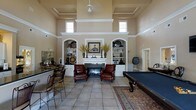Located at 100 Delaney Dr in Jacksonville, NC, this single-story space makes a striking impression with its grandly scaled great room, defined by soaring coffered ceilings and an abundance of natural light streaming in through clerestory windows positioned high along the walls. The room centers around a classic white fireplace flanked by symmetrical built-in arched shelving units, offering both decorative display space and functional storage with cabinetry below. Warm neutral tones throughout the space lend a welcoming, refined ambiance. A ceiling fan adds a practical touch while complementing the architectural elegance above. Large format tile flooring runs throughout, grounding the space with a clean, durable finish. To one side, a billiards table invites recreation, while a bar area with counter seating provides a social gathering spot. An area rug anchors the seating arrangement near the fireplace, where leather chairs create an intimate conversation area. The overall layout balances leisurely activity with relaxed comfort, making this a versatile and well-appointed communal space suited for both social events and everyday use.