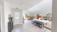Located at 1025 S Crater Rd in Petersburg, Virginia, this single-floor space offers a well-designed layout centered around a generous living room and connecting hallway. The living room immediately impresses with its dramatic vaulted ceilings and clerestory windows that flood the interior with abundant natural light throughout the day. Large front-facing windows further enhance the brightness while offering views of the surrounding greenery outside.

The flooring throughout is a light-toned hardwood-style material that complements the neutral, white and soft gray wall palette, creating a clean and airy atmosphere. A whitewashed brick accent wall adds a touch of texture and character to the living area, serving as a standout architectural feature. Crown molding along the ceiling transitions adds a refined, polished finish to the space.

The hallway connects seamlessly to a pass-through kitchenette area with white cabinetry and a compact countertop, providing functional utility without interrupting the open flow. High clerestory windows in this zone continue the theme of natural illumination.

Overall, the space carries a warm, inviting ambiance enhanced by high ceilings, plentiful natural light, and thoughtful architectural detailing — making it a bright and functional environment suited for a variety of uses.