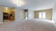 Located at 526 Middletown Rd in Hummelstown, Pennsylvania, this single-floor space offers a thoughtfully designed layout with a clean, neutral aesthetic throughout. The main living area is generously sized, featuring soft wall-to-wall carpeting in a warm beige tone that complements the light gray walls and bright white trim. An elegant chandelier anchors the living space, adding a touch of refinement to the open-concept design. Recessed lighting and a flush ceiling fixture provide additional illumination, ensuring the space remains bright and welcoming at any time of day. A set of classic French doors lines one wall, inviting natural light into the interior while offering potential access to an outdoor area. The adjacent kitchen is partially visible and features warm wood-toned cabinetry and a dark countertop, providing a functional and attractive cooking space. Additional doorways lead to further rooms, suggesting a practical and well-organized floor plan. Clean white baseboards and neutral walls tie the entire space together, creating a cohesive and move-in-ready environment. The overall ambiance is calm, open, and versatile, well-suited for a variety of living arrangements.