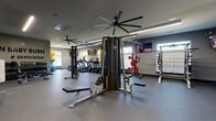 Located at 168 Headwater Circle in Mooresville, NC, this fitness facility is designed to support a full range of workout routines within a single-floor layout. The space is organized into distinct functional zones, allowing for both cardiovascular training and strength work without congestion.

The cardio section features a row of TRUE-brand treadmills and stair climbers positioned along one wall, accompanied by natural light from windows fitted with horizontal blinds. Mounted flat-screen televisions provide entertainment throughout the space. Large industrial-style ceiling fans are installed overhead, promoting airflow and comfort during workouts.

The strength training area includes a multi-station cable machine, a Smith machine, power racks, adjustable benches, and a dumbbell rack, offering versatility for a wide range of exercises. Rubber flooring covers the entire workout area, providing both durability and safety. Full-length mirrors along one wall allow users to monitor their form.

A motivational "Burn Baby Burn #Gymaholic" wall graphic adds personality to the space. An entry area with a water fountain and wall-mounted television connects the gym to an adjacent room visible through glass-paneled doors. Linear LED lighting runs along the ceiling, ensuring consistent, bright illumination throughout the facility.