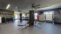 Located at 168 Headwater Circle in Mooresville, NC, this fitness facility is designed to support a full range of workout routines within a single-floor layout. The space is organized into distinct functional zones, allowing for both cardiovascular training and strength work without congestion.

The cardio section features a row of TRUE-brand treadmills and stair climbers positioned along one wall, accompanied by natural light from windows fitted with horizontal blinds. Mounted flat-screen televisions provide entertainment throughout the space. Large industrial-style ceiling fans are installed overhead, promoting airflow and comfort during workouts.

The strength training area includes a multi-station cable machine, a Smith machine, power racks, adjustable benches, and a dumbbell rack, offering versatility for a wide range of exercises. Rubber flooring covers the entire workout area, providing both durability and safety. Full-length mirrors along one wall allow users to monitor their form.

A motivational "Burn Baby Burn #Gymaholic" wall graphic adds personality to the space. An entry area with a water fountain and wall-mounted television connects the gym to an adjacent room visible through glass-paneled doors. Linear LED lighting runs along the ceiling, ensuring consistent, bright illumination throughout the facility.
