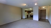 This single-floor space features an open-concept layout that connects a spacious carpeted living area to a well-equipped kitchen. The living area is finished with neutral-toned carpet and smooth walls, creating a clean and versatile space suitable for various furniture arrangements. Track lighting on the ceiling provides adjustable illumination throughout the main living space.

The kitchen is situated at the rear of the open layout and transitions from carpet to tile flooring. It features dark wood cabinetry offering ample storage, and is equipped with a full suite of appliances including a refrigerator, dishwasher, microwave, and range. A fluorescent overhead light fixture provides bright task lighting over the workspace. The kitchen's layout makes efficient use of its footprint with counter space along two walls.

A doorway visible from the main area hints at additional rooms, suggesting a functional floor plan beyond the open living and kitchen zone. The overall ambiance is practical and straightforward, with neutral finishes throughout that provide a blank canvas for personalization.