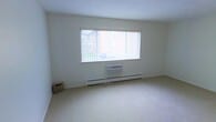 This single-floor space presents a clean, minimalist interior defined by neutral white walls and light-toned wall-to-wall carpeting that contribute to a bright, open atmosphere. The primary room visible features a large, wide picture window fitted with vertical blinds, allowing ample natural light to filter in while offering a view of the surrounding outdoor greenery. Below the window sits a wall-mounted air conditioning unit, providing climate control, complemented by a continuous baseboard heating system that runs along the base of the wall — together ensuring year-round comfort. The room's simple, uncluttered design emphasizes spaciousness, with smooth white ceilings and clean wall lines reinforcing a straightforward, functional aesthetic. A small woven basket in the corner adds a subtle organic touch to the otherwise pared-back space. The overall ambiance is calm and neutral, offering a practical and adaptable environment well-suited to a variety of uses. The single-floor layout ensures easy accessibility throughout, and the combination of natural light, efficient heating and cooling systems, and soft carpeted flooring creates a comfortable and livable setting.