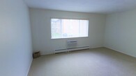 This single-floor space presents a clean, minimalist interior defined by neutral white walls and light-toned wall-to-wall carpeting that contribute to a bright, open atmosphere. The primary room visible features a large, wide picture window fitted with vertical blinds, allowing ample natural light to filter in while offering a view of the surrounding outdoor greenery. Below the window sits a wall-mounted air conditioning unit, providing climate control, complemented by a continuous baseboard heating system that runs along the base of the wall — together ensuring year-round comfort. The room's simple, uncluttered design emphasizes spaciousness, with smooth white ceilings and clean wall lines reinforcing a straightforward, functional aesthetic. A small woven basket in the corner adds a subtle organic touch to the otherwise pared-back space. The overall ambiance is calm and neutral, offering a practical and adaptable environment well-suited to a variety of uses. The single-floor layout ensures easy accessibility throughout, and the combination of natural light, efficient heating and cooling systems, and soft carpeted flooring creates a comfortable and livable setting.