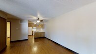 This single-floor space is designed with an open-concept layout that seamlessly connects the living area to the kitchen. The flooring throughout is a warm hardwood-style material, lending a cohesive and inviting feel to the interior. A ceiling fan with a built-in light fixture is centrally positioned, providing both air circulation and ambient lighting to the shared living and dining space.

The kitchen is defined by medium-toned oak cabinetry that runs along the upper and lower walls, offering generous storage. A peninsula with matching cabinetry extends into the open area, creating a natural division between the kitchen and living space while also serving as a convenient prep or casual dining surface. The kitchen is equipped with white appliances, including a refrigerator and a range with overhead ventilation. A sink is positioned along the counter, and a vent on the upper wall ensures proper air circulation.

The living area is notably open and bright, with clean white walls and a textured popcorn ceiling. Dark baseboards provide a grounding contrast to the light walls and warm floors, completing the space with a polished, functional aesthetic.