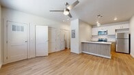 This efficiently designed single-floor unit offers a clean, modern aesthetic anchored by light wood-style flooring that runs continuously throughout the space, creating a cohesive and airy feel. The open-concept living area serves as the heart of the unit, featuring a ceiling fan with integrated lighting and a large window that allows natural light to fill the room. The entry door opens directly into this welcoming space, which flows naturally toward the kitchen and interior rooms.

The kitchen is well-appointed with a thoughtful two-tone cabinetry design, combining crisp white upper cabinets with warm wood-toned lower cabinets. A subway tile backsplash adds a classic touch, while the quartz peninsula countertop with an undermount sink provides both prep space and a natural divider from the living area. Stainless steel appliances — including a refrigerator, range, and over-the-range microwave — complete the functional and stylish setup.

The bathroom continues the modern design language with wood-toned floating vanity cabinetry, a white countertop, a rectangular wall-mounted mirror, and wood-look tile flooring. The space is clean and minimalist in presentation.

Neutral white walls throughout, recessed and pendant lighting, and consistent modern hardware tie the entire unit together into a cohesive, contemporary living environment.