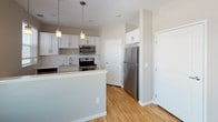 This single-floor unit offers a well-designed open layout that efficiently connects the main living and kitchen areas. The kitchen is a standout feature, outfitted with crisp white shaker cabinetry, a white subway tile backsplash, light stone countertops, and a full suite of stainless steel appliances including a range, over-the-range microwave, dishwasher, and refrigerator. A peninsula with pendant lighting above separates the kitchen from the living area, creating a natural gathering space while maintaining an open feel.

The living area benefits from large windows along one wall, allowing generous natural light to fill the space and providing views of the surrounding area. Light-toned hardwood flooring runs throughout the main living spaces, contributing to a clean, contemporary aesthetic against the neutral gray walls.

The bathroom is well-appointed with a granite-topped vanity, a large mirror with overhead lighting, a full soaking tub with shower, and hardwood-style flooring. Notably, in-unit laundry is conveniently located adjacent to the bathroom. The overall ambiance of the unit is modern and functional, with a cohesive neutral palette and quality finishes throughout.