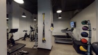 Located at 21 W Elm St in Chicago's Gold Coast neighborhood, this single-floor amenity space offers residents a well-appointed fitness center and a convenient on-site laundry facility. The gym area features an industrial-modern aesthetic, with exposed dark ceilings, structural concrete columns, and speckled epoxy flooring that lends a durable, professional feel throughout. The space is outfitted with a comprehensive range of equipment, including multiple treadmills equipped with individual screens, stationary bikes, an elliptical, and a rowing machine for cardio training. A dedicated strength zone features a full dumbbell rack with a weight bench, a cable machine, and a TRX suspension training system mounted to a central column. Medicine balls in various weights are neatly organized on a rack, adding versatility for functional training. Wall-mounted flat-screen televisions are positioned throughout the gym for entertainment. Pendant and recessed lighting illuminate the space effectively. Adjacent to the fitness area, the laundry room is clean and efficiently organized, featuring a row of front-loading washing machines and stacked dryers, all set on tiled flooring with a wall-mounted television for convenience. Together, these amenities provide a practical and functional environment for everyday resident needs.