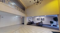 This expansive lobby presents an immediately impressive sense of scale, highlighted by dramatically soaring double-height ceilings that draw the eye upward to a decorative upper-level balcony. The balcony features ornate railings with detailed carved accents, adding an elegant architectural focal point to the space. A classic multi-arm chandelier hangs prominently overhead, complementing recessed lighting that illuminates the area evenly throughout.

The flooring consists of large-format light-toned ceramic tiles arranged in a geometric border pattern, lending a polished and formal character to the main floor. Dark runner mats define the primary circulation paths, guiding visitors from the entrance toward key functional areas. Entry points include glass-paneled doors with sidelights that allow natural light to filter in, and large potted trees flank these entryways, adding warmth and a natural element to the interior.

Functional amenities include a bank of individual mailboxes mounted along one wall and two elevator bays providing vertical access. A seating area along one side features upholstered chairs and a side table beneath a pair of framed landscape paintings, creating a comfortable waiting space. An additional seating arrangement with a small table and lamp offers further gathering potential. The warm yellow accent wall adds a welcoming, inviting ambiance throughout the lobby.