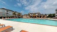 Located at 327 Guinevere Ln in Wilmington, North Carolina, this single-level outdoor amenity space serves as a vibrant community gathering hub designed for relaxation and recreation. At its centerpiece is a large, resort-style swimming pool with clear turquoise water, surrounded by a generously sized concrete sundeck lined with orange lounge chairs and umbrellas, offering ample space for sunbathing under the Carolina sky. The pool area is enclosed by a black wrought-iron fence with gated access, ensuring both safety and a clean aesthetic.

Adjacent to the pool, a covered pavilion provides a shaded retreat equipped with an outdoor kitchen featuring granite countertops, built-in grills, and seating at high-top tables with bright orange sling chairs — ideal for outdoor dining and social gatherings. Additional open-air seating areas with round tables are scattered throughout the patio, complementing the relaxed atmosphere.

Lush landscaping with tropical palm trees enhances the coastal feel of the space, while views of the surrounding multi-story residential buildings frame the area. The combination of functional amenities, thoughtful design, and vibrant furnishings creates an inviting outdoor environment that encourages community connection and leisure.
