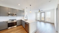 This thoughtfully designed single-floor unit offers a well-organized layout encompassing a kitchen, living room, bedroom, bathroom, office, and closet. Warm wood-look flooring flows consistently throughout, creating visual cohesion across the entire space.

The open-concept kitchen is a standout feature, equipped with full-height gray cabinetry, stainless steel appliances including a refrigerator, range, microwave, and dishwasher, and a white quartz peninsula with pendant lighting overhead. A white subway tile backsplash adds a clean, modern contrast to the gray cabinetry. The kitchen opens directly into the living area, which benefits from a ceiling fan and a glass door flanked by large windows that flood the space with natural light and offer exterior views.

The bedroom is a well-proportioned private retreat with dual windows providing additional natural light and outdoor views, complemented by a ceiling fan. It connects to a spacious walk-in closet outfitted with multi-tier wire shelving for generous storage.

The bathroom features an elongated dual-sink vanity with gray cabinetry, white quartz countertops, matte black fixtures, and large-format tile flooring. A dedicated office provides flexible space for work or additional use. A hallway with a double-door closet connects the living areas to the private rooms, completing a functional and modern floor plan.