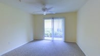 This single-floor space features a well-lit interior room finished with soft, neutral-toned carpet that extends throughout the floor, creating a comfortable and cohesive feel underfoot. The walls are painted in a light yellow hue, contributing to an open and airy atmosphere that is further enhanced by the abundance of natural light streaming in through a large sliding glass door. The door is dressed with vertical blinds, allowing for adjustable light control and privacy as needed. Beyond the glass, lush greenery is visible, providing a pleasant outdoor view that connects the interior to the surrounding landscape. A ceiling fan is centrally mounted overhead, offering both functional air circulation and a clean, understated design element. The room's simple, uncluttered layout allows for flexible furniture arrangement, making it adaptable to a variety of uses. The overall ambiance is calm and inviting, characterized by bright natural light, soft finishes, and a seamless visual connection to the outdoors through the sliding glass door.