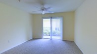 This single-floor space features a well-lit interior room finished with soft, neutral-toned carpet that extends throughout the floor, creating a comfortable and cohesive feel underfoot. The walls are painted in a light yellow hue, contributing to an open and airy atmosphere that is further enhanced by the abundance of natural light streaming in through a large sliding glass door. The door is dressed with vertical blinds, allowing for adjustable light control and privacy as needed. Beyond the glass, lush greenery is visible, providing a pleasant outdoor view that connects the interior to the surrounding landscape. A ceiling fan is centrally mounted overhead, offering both functional air circulation and a clean, understated design element. The room's simple, uncluttered layout allows for flexible furniture arrangement, making it adaptable to a variety of uses. The overall ambiance is calm and inviting, characterized by bright natural light, soft finishes, and a seamless visual connection to the outdoors through the sliding glass door.