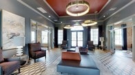 This elegantly designed lounge space makes an immediate impression with its soaring ceilings accented by a striking terracotta-red coffered ceiling detail at the center. Suspended from above are dramatic oversized brass ring pendant lights that serve as bold focal points while casting a warm, ambient glow throughout the room. The walls are finished in a cool slate gray, providing a sophisticated contrast to the warm tones of the furnishings and ceiling detail. Large, multi-pane windows and glass-paneled doors line the perimeter, flooding the space with abundant natural light and offering views to the outdoors. Dark, floor-length curtains frame each window, adding a layer of refinement and depth. The seating arrangement is thoughtfully curated, featuring a generous charcoal sectional sofa, a cognac leather ottoman, and several upholstered accent chairs positioned to encourage both conversation and relaxation. Underfoot, a geometric patterned area rug anchors the main seating area and adds visual interest to the hardwood flooring. A glass partition wall to one side allows for visual connectivity to an adjacent space while maintaining a sense of separation. The overall ambiance is upscale and inviting, blending contemporary design with classic architectural details.