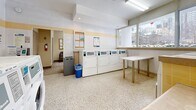 This shared laundry facility offers a clean, bright, and efficiently organized space designed to accommodate high-volume use. The main laundry area is generously proportioned, featuring multiple front-loading washers and stacked dryer units, all coin- or card-operated for convenient self-service. The layout is thoughtfully arranged, with dedicated folding tables positioned throughout the room to provide ample workspace for residents. A central island counter with a light wood finish adds both functionality and a touch of warmth to the otherwise utilitarian space.

The facility is finished with durable terrazzo flooring throughout the main area, providing a clean and easy-to-maintain surface. Walls are clad in white and yellow-accented tile, contributing to the bright and hygienic aesthetic. Large windows along multiple walls flood the space with natural light and offer views of the surrounding urban streetscape.

An adjacent waiting area provides a comfortable place to relax while laundry cycles run, furnished with seating, a wall-mounted television, and a bulletin board for community notices. Recessed and fluorescent lighting ensures the entire facility is well-lit at all times. Waste and recycling receptacles are conveniently placed throughout. The space is clearly maintained to a high standard, making it a practical and welcoming amenity.