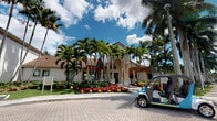 This single-story building presents a striking Mediterranean-inspired architectural design, characterized by a terracotta barrel-tile roof, stucco exterior walls, and elegant arched entryways framed by decorative columns. The grand front entrance is approached via a paved brick circular drive, adding to the sense of arrival and prestige. Tall, majestic royal palms line the property, complementing the vibrant tropical landscaping that includes colorful flowering beds of red and yellow blooms, manicured hedges, and lush green lawns. Stone accent details on the facade add texture and visual interest, while large windows allow natural light to fill the interior. The overall ambiance is that of a polished, resort-style facility — warm and welcoming, with a distinctly South Florida character. The well-maintained grounds and thoughtfully designed exterior reflect a commitment to curb appeal and a high standard of presentation. The combination of classical architectural elements and tropical surroundings creates an inviting, upscale atmosphere that is both functional and aesthetically distinguished.