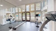 This fitness center is thoughtfully designed to support a well-rounded workout experience. The space is divided into distinct functional zones, each serving a specific training purpose. A standout architectural feature is the cardio area's soaring ceiling height, framed by two tiers of white-trimmed, multi-pane windows and glass-paneled French doors that flood the room with natural light and offer views of the surrounding greenery outside. The reflective quality of full-wall mirrors further enhances the sense of openness throughout this section. Durable rubber flooring provides a practical and safe surface underfoot.

The cardio zone is equipped with treadmills, stationary bikes, and an elliptical trainer, while a bold blue accent wall adds a vibrant, energizing visual element to this area. The strength training section features cable resistance machines and a weight bench arranged across a spacious open floor, with additional full-wall mirrors allowing users to monitor their form. Recessed lighting in the textured ceiling illuminates this zone effectively. Together, the two areas create a cohesive, well-lit, and fully functional fitness environment suited for a comprehensive range of exercise routines.