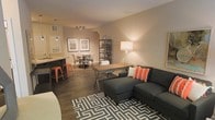 This well-appointed single-floor residence offers an open-concept design that seamlessly connects the living, dining, and kitchen areas into one cohesive and inviting space. The living area serves as the centerpiece, anchored by a plush dark sectional sofa adorned with bold orange accent pillows and set atop a striking geometric black-and-white area rug. A large floor lamp provides warm ambient lighting, complementing the overall cozy atmosphere. Adjacent to the living area, a versatile workspace features an industrial-style metal desk paired with a metal shelving unit, ideal for both work and display. The dining area is defined by a round table surrounded by classic chairs, with framed artwork on the warm-toned walls adding a curated, personal touch. A pendant light fixture overhead enhances the intimate feel of the dining space. The kitchen flows naturally from the main living area and is highlighted by a breakfast bar with vibrant orange bar stools that add a pop of color and a casual dining option. Dark hardwood-style flooring runs throughout, grounding the space and tying together the modern industrial aesthetic. Neutral wall tones and warm lighting create an inviting ambiance throughout.