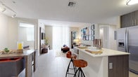 This contemporary single-floor residence showcases a thoughtfully designed open-concept layout that seamlessly connects the kitchen and living areas. The kitchen is outfitted with dark wood-grain cabinetry, a gray subway tile backsplash, and a full suite of stainless steel appliances, including a side-by-side refrigerator with water dispenser, electric range, built-in microwave, and dishwasher. A peninsula with a crisp white countertop provides seating for two and serves as a natural divider between the kitchen and the main living space. Large-format light tile flooring runs throughout the entry and kitchen areas, transitioning into wood-look flooring in the living room, which is bathed in natural light from expansive floor-to-ceiling windows dressed with vertical blinds. The bedroom offers plush carpeting, ample space, and direct access to a spacious walk-in closet featuring wire shelving. The bathroom is finished with large-format gray tile flooring, a double-sink vanity with dark cabinetry and white countertops, a wide mirror with overhead lighting, and a glass-enclosed shower. The neutral color palette and modern finishes throughout lend the space a clean, cohesive aesthetic.