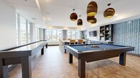 This well-appointed amenity lounge is designed to offer a dynamic and social atmosphere across a single floor. The open-plan layout is anchored by a games area, which features two full-size pool tables with blue felt surfaces and a shuffleboard table, all positioned beneath a striking cluster of decorative bronze pendant lights that add warmth and visual interest to the space.

Adjacent to the games area, a comfortable lounge zone is furnished with tufted leather armchairs, natural wood side tables, and layered area rugs atop a geometric parquet-patterned tile floor. A wall-mounted flat-screen TV is flanked by framed artwork and adjustable sconce lighting, creating an inviting setting for relaxed gatherings.

A dining area with a high-top table and bar-style seating is positioned near large windows that allow generous natural light to fill the interior. Nearby, a built-in kitchenette with open shelving and a sink provides functional convenience for entertaining.

A bright sunroom serves as a transitional space between the interior and outdoors, enclosed by floor-to-ceiling windows and glass doors, and equipped with a ceiling fan and casual seating. This area offers panoramic outdoor views, enhancing the overall airy and welcoming character of the lounge.