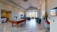 This well-appointed single-floor community clubhouse offers a thoughtfully designed gathering space with a variety of distinct areas to relax, socialize, and unwind. The entry lobby sets an inviting tone with soaring high ceilings, soft wall sconces, large glass-paned doors, and a built-in bench with a bold red cushion that adds a pop of color against the light gray walls and wood-look flooring found throughout.

The main lounge area is the centerpiece of the space, featuring a soaring vaulted ceiling with dual ceiling fans, a classic billiards table, and multiple seating arrangements. A textured accent wall frames a sleek modern fireplace flanked by stylish teal accent chairs, while a wall-mounted television adds entertainment value. Large bay windows with teal trim flood the adjacent sitting area with natural light and offer pleasant outdoor views, complemented by an assortment of contemporary seating options.

A cozy nook near the entry features a high-top round table with industrial-style stools, a decorative mirror, and wall-mounted television — ideal for casual conversation. The kitchenette is equipped with white cabinetry, dark countertops, a stainless steel refrigerator, a sink, and a mounted television, providing all the essentials for hosting gatherings. Throughout, the consistent wood-look flooring, soft neutral palette, and modern furnishings create a cohesive and welcoming ambiance.