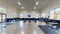 The Canopy Living fitness center at 4400 SW 20th Ave in Gainesville, Florida, is a well-appointed, single-floor facility designed to support a wide range of fitness activities. The space is defined by soaring ceilings and a clean, modern aesthetic, with crisp white walls accented by a bold navy blue wainscoting that runs throughout, creating a cohesive and energetic atmosphere.

The main cardio area is expansive and open, featuring polished concrete floors that reflect the bright overhead lighting. This zone is equipped with a row of treadmills, ellipticals, stair climbers, and stationary cycling bikes, all arranged to allow ample space between each machine. A wall-mounted flat-screen television provides entertainment during workouts, while large windows on multiple walls bring in natural light and views of the surrounding landscape.

Adjacent to the cardio floor is a dedicated strength and functional training area, outfitted with rubber flooring, free weights, barbells, medicine balls, rowing machines, and a variety of benches and workout mats. This zone benefits from the same high ceilings and generous natural light.

A standout feature of the facility is its full-height rock climbing wall, adding a unique and dynamic element to the fitness offerings. An open activity area with padded mats provides space for yoga, stretching, or group fitness. The "Go Gators" spirit wall reinforces the community's connection to Gainesville's vibrant collegiate culture.