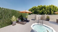 This rooftop terrace offers a well-appointed open-air retreat designed for relaxation and outdoor living. The spacious deck is finished in a clean, neutral concrete surface and is divided into several distinct functional zones, each thoughtfully furnished for comfort and leisure.

At the center of the terrace is a built-in hot tub, complete with handrail entry and active jets, serving as the focal point of the outdoor space. Adjacent to the spa, a pair of wicker chaise lounges with cushions sits beneath a large shade umbrella, ideal for sunbathing or unwinding. Tall snake plants in sleek dark planters are strategically placed throughout, adding vertical greenery and a contemporary aesthetic.

A dedicated lounge area features a wicker sectional sofa with plush white cushions, arranged around a rustic wood-top coffee table, all sheltered under another generous patio umbrella. A separate wicker dining set with chairs provides an additional gathering spot for outdoor meals or casual conversation.

Privacy walls of textured stucco surround the perimeter, while a green faux-hedge panel adds a lush, garden-like boundary on one side. Mature trees visible beyond the walls contribute a natural canopy backdrop. Views of neighboring rooftops and distant buildings give a sense of urban elevation. The terrace is accessed via a stairwell with a clearly marked exit, making it both accessible and practical.
