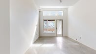 Located at 5460 N Interstate Ave in Portland's vibrant North Interstate corridor, this efficiently designed single-floor unit offers a clean, contemporary aesthetic throughout. The living room serves as the centerpiece of the space, characterized by soaring ceilings that amplify the sense of openness well beyond its compact footprint. Large sliding windows paired with a transom window above flood the room with natural light, while a glass-paneled entry door provides direct street access and an urban view that reflects the neighborhood's lively character. Polished concrete floors run continuously throughout the unit, lending an industrial-modern edge and a seamless visual flow between spaces. The kitchen is tucked neatly behind a partial wall partition, featuring white cabinetry, pendant lighting, and white appliances that maintain the unit's clean, minimalist palette. The bathroom is thoughtfully appointed with a single-basin vanity, standard fixtures, and concrete flooring consistent with the rest of the unit, along with a closet area that adds practical storage. Crisp white walls throughout create a bright, gallery-like ambiance, making the space feel fresh and versatile. This unit is well-suited for those seeking a modern, low-maintenance urban residence in North Portland.