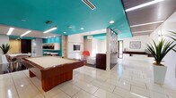 Located at 4940 Paso Robles Ave in the Encino neighborhood of Los Angeles, this single-floor amenity space combines modern design with a lively, mid-century-inspired aesthetic. The space is immediately distinguished by its striking teal-painted ceilings, which contrast boldly against crisp white walls and create a cohesive visual identity throughout.

The main gathering area is generously proportioned and centers around a full-size wood-framed pool table set atop large-format, polished white tile flooring that reflects light and enhances the open feel of the room. Recessed lighting and linear fluorescent fixtures illuminate the space evenly, while a decorative pendant light adds a touch of warmth to the lounge nook. The lounge area features a wall-mounted flat-screen TV, an accent chair in vivid orange-red, and white seating, framed by a textured stone column that adds an organic architectural element.

A stylish corridor connects the main area to additional spaces, featuring an orange upholstered bench, nature-inspired wall art, and a glass display case with teal interior shelving. The kitchenette is well-appointed with white cabinetry, a stainless steel sink, hexagonal tile backsplash, compact refrigerator, and coffee station, complemented by rich dark tile flooring and a woven roller shade. An adjacent dining area with striped chairs and a white table, alongside a tall potted plant, adds a casual and welcoming touch. A decorative geometric room divider and views to the outdoors complete the inviting atmosphere.