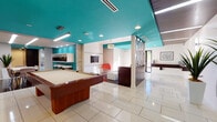 Located at 4940 Paso Robles Ave in the Encino neighborhood of Los Angeles, this single-floor amenity space combines modern design with a lively, mid-century-inspired aesthetic. The space is immediately distinguished by its striking teal-painted ceilings, which contrast boldly against crisp white walls and create a cohesive visual identity throughout.

The main gathering area is generously proportioned and centers around a full-size wood-framed pool table set atop large-format, polished white tile flooring that reflects light and enhances the open feel of the room. Recessed lighting and linear fluorescent fixtures illuminate the space evenly, while a decorative pendant light adds a touch of warmth to the lounge nook. The lounge area features a wall-mounted flat-screen TV, an accent chair in vivid orange-red, and white seating, framed by a textured stone column that adds an organic architectural element.

A stylish corridor connects the main area to additional spaces, featuring an orange upholstered bench, nature-inspired wall art, and a glass display case with teal interior shelving. The kitchenette is well-appointed with white cabinetry, a stainless steel sink, hexagonal tile backsplash, compact refrigerator, and coffee station, complemented by rich dark tile flooring and a woven roller shade. An adjacent dining area with striped chairs and a white table, alongside a tall potted plant, adds a casual and welcoming touch. A decorative geometric room divider and views to the outdoors complete the inviting atmosphere.