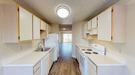Located at 7024 North Colton Street in Spokane, Washington, this single-floor unit offers a practical and well-organized layout. The galley-style kitchen is designed for efficiency, featuring white cabinetry with wood-tone trim, white appliances including an electric coil range and refrigerator, a built-in dishwasher, a stainless steel sink, and an over-range ventilation hood. Ample upper and lower cabinet storage lines both sides of the kitchen, and a window at the far end brings in natural light. The kitchen opens directly into the dining area and flows seamlessly into the spacious living room, which is highlighted by rich hardwood-style flooring, neutral warm-toned walls, and a sliding glass door that fills the space with natural light. The bedroom features the same hardwood-style flooring, a large window, a baseboard heating unit, and access to a walk-in closet. The bathroom includes a wide vanity with a large mirror, multi-bulb lighting fixture, generous counter space with white cabinetry below, and a full tub-shower combination with a curved curtain rod. Textured ceilings are present throughout the unit, contributing to a consistent finish. The overall design is clean, functional, and well-lit, making for a comfortable living environment.