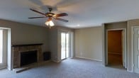 This single-floor living space offers a comfortable, functional layout centered around a classic brick fireplace with a dark mantel, serving as the room's focal point. The fireplace adds a traditional character to the space and provides a natural gathering area. Neutral-toned carpeting extends throughout the room, contributing to a warm, understated aesthetic that pairs well with the beige walls.

A ceiling fan with a built-in light fixture is centrally mounted, providing both air circulation and ambient lighting. Recessed lighting supplements the overhead illumination, ensuring the space is well-lit. A sliding glass door along one wall fills the room with natural light and opens to a balcony, offering a view of the outdoors and providing easy access to fresh air.

The open layout allows for flexible furniture arrangements, making the space adaptable to various living needs. Clean white trim and baseboards frame the room neatly, while a doorway visible on the right side suggests additional rooms beyond. Overall, the space presents a practical and inviting environment suitable for everyday living.