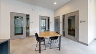 Located at 18905 E 59th Ave in Denver, CO, this contemporary single-floor communal space is thoughtfully designed to serve both social and professional needs. The kitchen and dining area form the heart of the space, anchored by a sleek, large-format island with a dark stone countertop and lined with upholstered bar seating. Overhead, stylish black pendant lights with brass accents cast a warm glow over the space. The kitchen wall features a striking combination of white and deep green subway tile as a backdrop, framing built-in flat-screen displays and white cabinetry outfitted with stainless steel appliances, including a dishwasher and microwave. Polished concrete flooring flows seamlessly throughout, lending a clean, industrial aesthetic. Adjacent to the kitchen, a casual lounge area offers comfortable seating with bold orange armchairs set beneath large windows that invite natural light. A wooden dining table with black chairs provides an additional gathering spot. Multiple private work pods line the perimeter, each enclosed by full-length glass-paneled gray doors, offering quiet, focused workspaces visible from the main area. Soaring ceilings and a neutral white palette enhance the sense of openness, creating a versatile environment well-suited for collaboration, dining, and focused work.