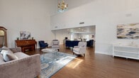 The Bell Oaks Apartments Lounge in Montgomery, Alabama, is a generously scaled single-floor community gathering space defined by its striking architectural character and thoughtfully arranged interiors. The most prominent feature of the main entry area is its dramatically vaulted ceiling, accented by exposed beams and adorned with elegant brass chandeliers that lend a refined, classic ambiance. Horizontal shiplap paneling along the upper walls adds textural interest, while white wainscoting throughout maintains a clean, polished aesthetic.

Rich hardwood flooring runs throughout the space, anchoring multiple distinct seating zones. The front lounge area features tufted accent chairs and navy blue upholstered sofas arranged around glass-top coffee tables atop patterned area rugs, creating an inviting setting for casual conversation. Deeper into the lounge, a fireplace framed in white brick serves as a focal point for another comfortable seating arrangement. A built-in bench flanks the fireplace, adding functional charm.

An expansive open area with a dropped tile ceiling provides flexible event space, flowing toward a kitchenette equipped with counter space and appliances. Large glass-paneled doors along the perimeter allow natural light to fill the space while offering views to the outdoors, enhancing the overall warmth and openness of this well-appointed community lounge.