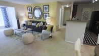 This single-floor residence offers a thoughtfully designed open-concept layout that blends comfort and style. The living area serves as the heart of the home, featuring warm yellow accent walls that lend a cheerful, inviting ambiance throughout. Plush wall-to-wall carpeting adds warmth underfoot, while the generous floor space allows for flexible and comfortable furniture arrangements. The living room is highlighted by a stylish gallery wall arrangement featuring framed artwork and a decorative round mirror, creating an eye-catching focal point. A dark upholstered sectional sofa, accent chair, and coordinating area rug complete the cohesive, contemporary look. The space flows naturally into a hallway corridor that connects to additional rooms, maintaining an open and airy feel. To one side, a kitchen area is partially visible, featuring a raised breakfast bar countertop with stone-look surfaces, offering a casual dining and entertaining option. Ceiling-mounted lighting fixtures provide consistent illumination throughout. The overall design palette of yellows, grays, and neutral tones creates a balanced and modern aesthetic, making this a comfortable and functional single-level living space.