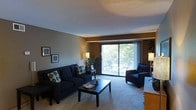 This single-floor space centers around a welcoming living room that balances comfort and functionality. The room is anchored by a large sliding glass door that floods the space with natural light and offers a lush, tree-lined outdoor view, creating a refreshing connection to the exterior. A private balcony is accessible through the sliding door, extending the living area outdoors and providing a pleasant spot for relaxation.

The living room is furnished with a deep navy blue sofa and a matching armchair, both accented with decorative throw pillows, lending a cohesive and inviting aesthetic. A dark rectangular coffee table sits at the center, complementing the seating arrangement. Soft wall-to-wall carpeting adds warmth underfoot and contributes to the room's cozy atmosphere.

Warm ambient lighting from floor lamps and wall-mounted sconces enhances the neutral-toned walls, creating a calm and comfortable environment throughout the day and evening. Framed wall art adds a personal touch to the space. The overall ambiance is relaxed and livable, with thoughtful details that make the space feel both functional and homey — ideal for everyday living and casual entertaining.