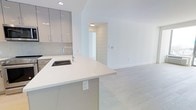 This single-floor space showcases a clean, contemporary design with an open-concept layout that connects the kitchen and living areas in a seamless, functional arrangement. The kitchen is appointed with high-gloss, light grey cabinetry featuring slim bar-pull hardware, paired with a crisp white countertop that extends into a peninsula with an integrated undermount sink and a polished chrome faucet. A subtle tile backsplash adds texture behind the cabinetry, complementing the overall neutral palette. Recessed lighting provides focused illumination throughout. The open living area extends naturally from the kitchen, bathed in bright, even light that enhances the airy atmosphere. Light-toned wide-plank hardwood flooring runs continuously throughout the space, adding warmth while maintaining the minimalist aesthetic. The white walls and clean architectural lines contribute to the sense of openness and spaciousness. A doorway visible from the main area leads to an additional room, suggesting a well-organized floor plan. Overall, the space presents a bright, modern environment with quality finishes and a cohesive, neutral design scheme.