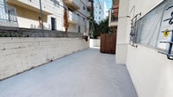 This single-level outdoor space presents a functional and characterful courtyard enclosed by painted concrete block walls, offering a well-defined and private environment. The smooth concrete flooring extends throughout, providing a clean, low-maintenance surface across the full footprint of the space.

A prominent wooden gate serves as the primary entry point, adding a warm, natural contrast against the light-toned masonry walls. An exterior staircase with black metal railings is integrated into the building facade, connecting the courtyard to the upper levels of the adjoining structure. The building exterior is finished in a neutral stucco, complemented by decorative iron window grilles that add an architectural detail to the facade.

Greenery plays a notable role in the ambiance of this space. Climbing vines and ivy grow along the walls and adjacent fencing, softening the hardscape and introducing a lush, organic quality to the environment. A dedicated planter ledge along the brick wall displays an assortment of potted plants, contributing further to the courtyard's natural character.

The space benefits from open-sky exposure, allowing for ample natural light throughout the day. Surrounding multi-story residential buildings and mature trees, including a visible palm, frame the outdoor area and reflect its urban setting.