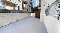 This single-level outdoor space presents a functional and characterful courtyard enclosed by painted concrete block walls, offering a well-defined and private environment. The smooth concrete flooring extends throughout, providing a clean, low-maintenance surface across the full footprint of the space.

A prominent wooden gate serves as the primary entry point, adding a warm, natural contrast against the light-toned masonry walls. An exterior staircase with black metal railings is integrated into the building facade, connecting the courtyard to the upper levels of the adjoining structure. The building exterior is finished in a neutral stucco, complemented by decorative iron window grilles that add an architectural detail to the facade.

Greenery plays a notable role in the ambiance of this space. Climbing vines and ivy grow along the walls and adjacent fencing, softening the hardscape and introducing a lush, organic quality to the environment. A dedicated planter ledge along the brick wall displays an assortment of potted plants, contributing further to the courtyard's natural character.

The space benefits from open-sky exposure, allowing for ample natural light throughout the day. Surrounding multi-story residential buildings and mature trees, including a visible palm, frame the outdoor area and reflect its urban setting.
