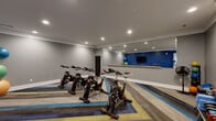 Located at 7029 W Friendly Ave in Greensboro, NC, this dedicated exercise room offers a focused and functional fitness environment. The space is thoughtfully designed with a bold color palette, featuring gray walls accented by a striking cobalt blue feature wall that adds energy and visual depth to the room.

The flooring is a standout element — a multi-toned striped carpet in shades of blue, teal, yellow, and gray lends a dynamic, modern aesthetic throughout. Recessed lighting in the smooth white ceiling provides consistent, even illumination across the entire space.

A row of spin bikes serves as the centerpiece of the room, catering to cycling workouts and group fitness sessions. Along one wall, a large floor-to-ceiling mirror is paired with a wooden ballet barre, making the space suitable for stretching, barre workouts, or dance-based exercise. A mounted flat-screen television adds a multimedia element for guided workouts or entertainment.

A dedicated free weights and accessories station lines another wall, stocked with kettlebells, dumbbells, resistance equipment, and stability balls, offering a well-rounded range of strength and functional training options. A coat hook station near the entrance provides convenient storage upon arrival.

The room is accessed through a clean white-trimmed doorway that connects to a stairwell, integrating the fitness space seamlessly into the broader building layout.