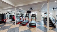 This two-story fitness facility offers a comprehensive workout environment designed to accommodate a wide range of exercise needs. The space is characterized by a bold checkerboard-patterned floor in contrasting slate and tan tones, which flows consistently throughout both levels and contributes to the facility's clean, polished aesthetic. Crisp white walls, recessed lighting, and ceiling fans maintain a bright and comfortable atmosphere, while large double-hung windows on multiple sides invite abundant natural light and views of the surrounding exterior.

The main floor houses an extensive selection of cardio equipment, including treadmills, stair climbers, stationary bikes, rowing machines, and ellipticals, all arranged across a generously open layout. A dedicated functional training room features stability balls, resistance bands, medicine balls, and foam rollers stored on organized racks. Mirrored walls in select areas enhance the sense of space and support form-focused training. Wall-mounted televisions are positioned throughout for convenience.

A sleek staircase with a modern black horizontal-rail railing connects the two floors. The upper level continues the same flooring and design language, featuring additional equipment zones and framed artwork. A dual water fountain and bottle-filling station is conveniently located on-site. The overall design balances functionality with a refined, welcoming ambiance.
