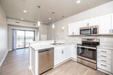 This single-floor unit presents a clean, contemporary design with a thoughtful open layout. The kitchen serves as a central feature, outfitted with white shaker cabinetry, light marble-style countertops, and a full suite of stainless steel appliances including a refrigerator, electric range, microwave, and dishwasher. A peninsula with an undermount sink and pendant lighting above creates a natural divide between the kitchen and the living area, which flows toward a sliding glass door opening onto a private balcony with open outdoor views.

The living space is finished with light wood-look flooring, lending warmth and continuity throughout the main area. The bedrooms feature soft carpeting and large windows fitted with horizontal blinds, allowing for ample natural light while maintaining privacy. A ceiling fan is present in one bedroom for added comfort.

The bathroom is well-appointed with a wide white vanity, quartz-style countertop, and black hardware accents paired with a large mirror. Conveniently accessible from the bathroom area is an in-unit stacked washer and dryer, adding practical functionality. The unit's neutral color palette, recessed lighting, and modern finishes throughout contribute to a bright, move-in-ready atmosphere.
