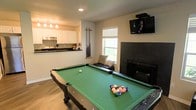 This single-floor space is thoughtfully designed for entertainment and relaxation, combining a recreational area with functional living amenities. The centerpiece of the main room is a full-sized pool table with a green felt surface, positioned to allow comfortable play from all sides. A wall-mounted flat-screen TV is installed in the corner, providing an excellent viewing angle from anywhere in the room. Adjacent to the TV, a dark-tiled fireplace adds a warm, inviting focal point to the space, complemented by a cozy seating bench with decorative pillows positioned nearby. A wall-mounted cue rack keeps billiard accessories neatly organized and within reach. Natural light filters in through a window overlooking what appears to be a green outdoor area, brightening the room alongside recessed lighting overhead. The flooring throughout is a light-toned wood laminate, lending a clean and modern aesthetic. An open-concept kitchen is visible from the main area, featuring white cabinetry, dark countertops, pendant lighting, and a gas range — keeping the host connected to guests during gatherings. The overall ambiance is casual, functional, and well-suited for both everyday use and social entertainment.