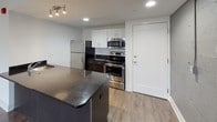 Located at 3075 W Grand Blvd in Detroit, Michigan, this single-floor unit offers a thoughtfully designed layout with modern finishes throughout. Wood-look luxury vinyl flooring runs continuously across all rooms, providing a cohesive and contemporary aesthetic.

The kitchen is a standout feature, equipped with stainless steel appliances including a refrigerator, range, and over-the-range microwave. White upper cabinetry contrasts with dark lower cabinets and a dark stone countertop island, creating a sleek, functional workspace. Recessed and track lighting illuminate the space effectively.

A long hallway connects the main living areas to the private rooms, with crisp white walls and subtle wall-mounted lighting guiding the way. The main living area is spacious and open, featuring an exposed painted concrete block accent wall that adds an industrial character, complemented by a window offering views of the surrounding Detroit cityscape.

Additional rooms provide flexible spaces with neutral white walls, windows with city views, and built-in ventilation. The bathroom is clean and functional, finished with white tile flooring, a tub and shower combination with a grab bar, and gray accent walls. Throughout the unit, the design balances urban industrial elements with modern, clean finishes to create a practical and stylish living environment.