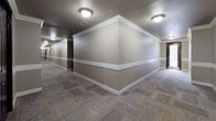 This community hallway presents a thoughtfully designed shared corridor that balances functionality with refined aesthetics. The hallway extends across a single floor, offering residents a well-maintained path connecting individual units, a stairwell, and elevator access.

The walls are finished in a warm taupe-gray tone, accented by crisp white crown molding along the ceiling line and a matching white chair rail running horizontally at mid-wall height, lending the corridor a classic, structured appearance. The textured ceiling adds subtle depth, while flush-mount ceiling light fixtures provide consistent, even illumination throughout the length of the passage.

Underfoot, a patterned carpet in muted earth tones features a geometric tile-like arrangement that adds visual interest without overwhelming the space. Dark-finished unit doors with gold numbering and hardware are evenly spaced along both sides of the hallway, creating a rhythmic and orderly presentation.

A dedicated elevator alcove branches off the main corridor, featuring tile flooring and the same cohesive wall treatment. Emergency exit signage and a fire safety panel are discreetly integrated near the stairwell entrance, ensuring safety features are accessible without detracting from the overall design. The corridor's layout is efficient, clean, and welcoming.
