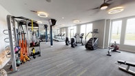 Located at 6137 Telluride Way in Denver, Colorado, this single-floor fitness center is a thoughtfully designed, contemporary facility built to support a wide range of workout routines. The space is expansive and open, finished with durable patterned carpet flooring and neutral gray walls that create a clean, professional atmosphere. Large windows line the perimeter, flooding the facility with natural light while offering views of the surrounding open landscape — an inviting backdrop for cardio workouts. A glass door provides direct access to an outdoor area, further connecting the interior to the outdoors. The cardio zone is well-stocked with treadmills, ellipticals, stair climbers, and a rowing machine, all positioned to take advantage of the scenic window views. A strength training section features a comprehensive dumbbell rack with floor-to-ceiling mirrors, weight benches, and cable machines. Circular halo light fixtures and ceiling fans provide functional and ambient lighting throughout. A standout artistic mural depicting an active outdoor scene adds visual character to the strength area. A glass-enclosed studio room offers a private space for stretching, balance training, or small group workouts, complete with stability balls and additional accessories. Built-in shelving and organized equipment storage round out this fully functional fitness facility.