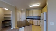 This single-floor residential space is thoughtfully designed to maximize functionality and comfort. The kitchen serves as a central highlight, featuring light wood cabinetry that extends to the ceiling for generous storage capacity. Dark laminate countertops provide a contrasting visual anchor, while a built-in sink and full suite of appliances — including a refrigerator, dishwasher, and range — ensure a fully equipped cooking environment. A peninsula with matching dark countertop extends into the adjoining area, creating a natural transition between the kitchen and living space while offering additional workspace and casual seating potential. The flooring transitions from light vinyl in the kitchen to carpet in the adjacent area, subtly defining each zone. Crown molding runs along the upper walls, adding a refined architectural detail throughout. Recessed and overhead lighting illuminate the space effectively. Two sets of closet doors are visible along one wall, offering practical storage solutions. The overall ambiance is clean, neutral, and move-in ready, with a layout that balances open-concept living with defined functional areas.