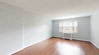 This single-floor space offers a well-organized layout with a consistent design language defined by soft blue-gray walls, warm hardwood flooring, and bright white trim throughout. The living area is generously proportioned, anchored by a bank of multi-pane windows that admit abundant natural light and offer views of the surrounding brick exterior. A white radiator cover beneath the windows adds a classic architectural detail.

The kitchen is efficiently designed with white cabinetry, granite countertops, white subway tile backsplash, and stainless steel appliances including a gas range and over-the-range microwave. A window above the counter allows natural light to fill the space, creating a pleasant cooking environment.

The bedrooms continue the hardwood flooring theme. One bedroom features dual windows with radiator heating, providing both light and warmth. Another bedroom includes a wall-mounted ductless split HVAC unit for supplemental climate control, along with closet access and direct hallway connectivity. Closet space is available with open-rod configurations.

The bathroom maintains a clean, classic aesthetic with white square wall tiles, a full bathtub with hand-held shower, chrome fixtures, and a window overlooking greenery. The overall ambiance of the space is calm, neutral, and move-in ready.